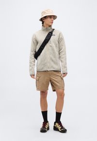 Νεαρός άνδρας που φοράει μπεζ καπέλο τύπου bucket, ανοιχτό γκρι φλις με φερμουάρ, ταμπά cargo σορτς, μαύρη τσάντα ώμου, μαύρες κάλτσες και μπεζ παπούτσια πεζοπορίας, όρθιος.