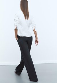 Blouse blanche à manches longues avec des fentes aux poignets, associée à un pantalon large à côte noir. La mannequin marche, vue de dos.