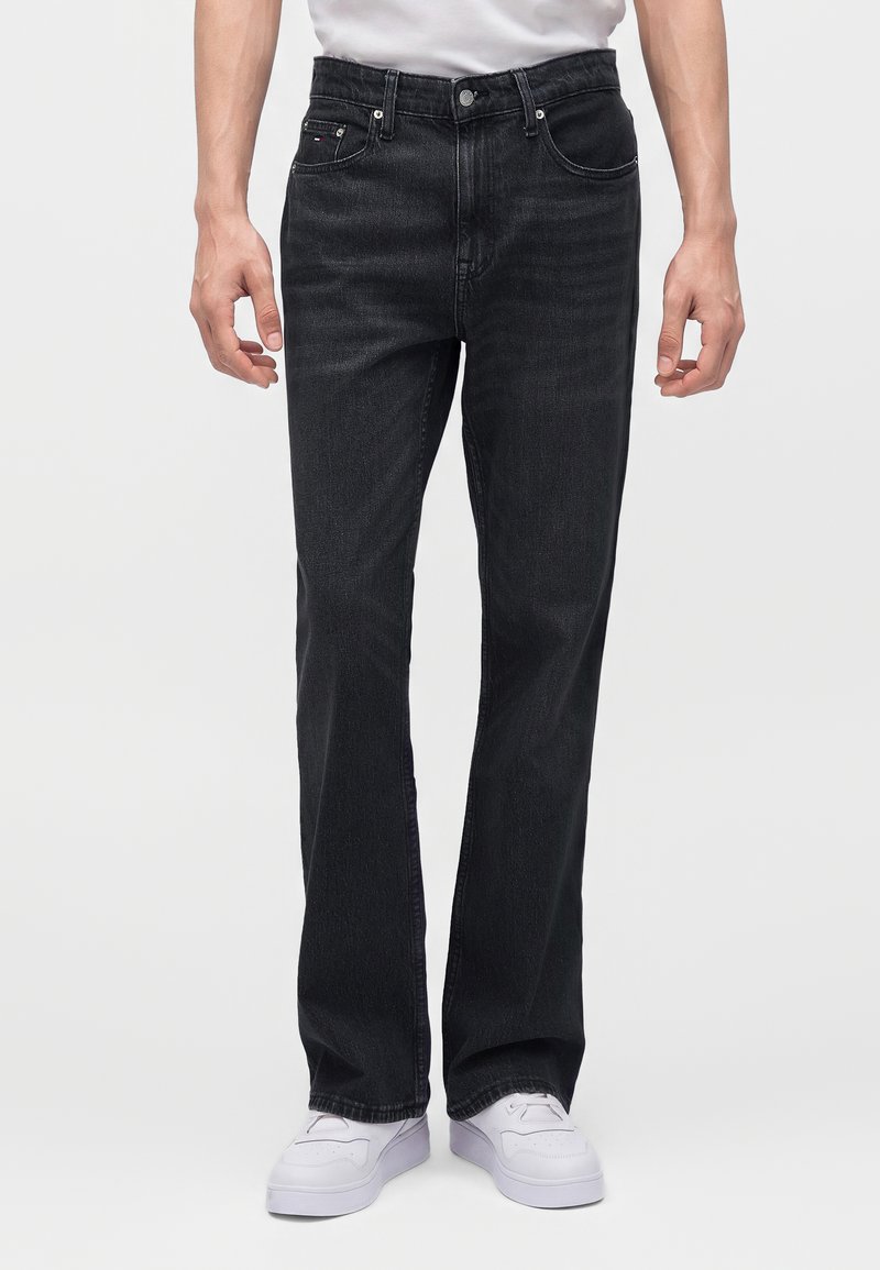 Homme portant un jean noir droit et des baskets blanches, debout devant un fond clair uni.