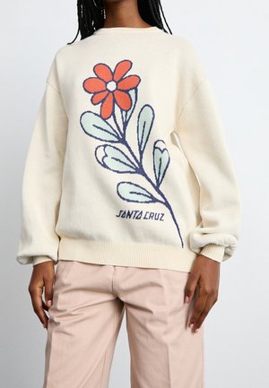 Personne portant un pull crème avec un grand motif de fleur rouge et de feuilles vertes et le texte "Santa Cruz" à l’avant.