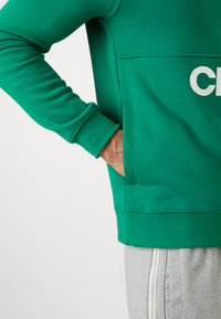 Sweatshirt en coton vert avec une coupe décontractée, featuring une poche avant et une lettrage audacieux en blanc. Associé à un pantalon de survêtement gris avec des fermetures éclair.