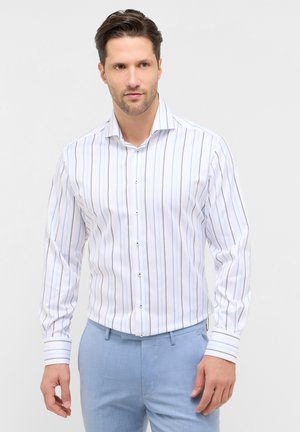 Uomo che indossa una camicia bianca con sottili righe verticali blu e pantaloni azzurri, in piedi contro uno sfondo semplice.