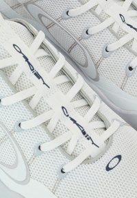 Weiße Mesh-Sportschuhe mit hellgrauen Akzenten, flachen Schnürsenkeln und einer strukturierten Grip-Sohle, mit Branding an den Seiten und der Zunge.
