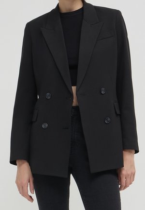 Frau trägt einen schwarzen, doppelt geknöpften Blazer über einem schwarzen Croptop und schwarzen Jeans, steht vor einem schlichten hellen Hintergrund.