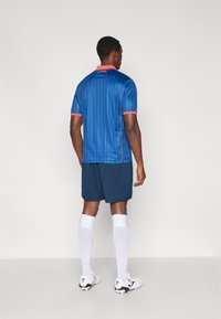 Kék sportmez függőleges vékony csíkokkal, piros gallérral és ujjakaszalmakkal, sötétkék rövidnadrággal és fehér zoknival párosítva. A mez hátoldalán logó található.