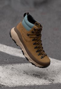 Stivale da hiking Columbia marrone e teal con suola spessa e lacci neri, sospeso sopra l'asfalto con segnature stradali bianche.