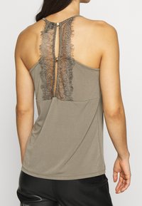 Grå ärmlös topp med en spetspanel på baksidan, som har ett nyckelhål och guldknappar vid halsen. Mjuk, slät textur.