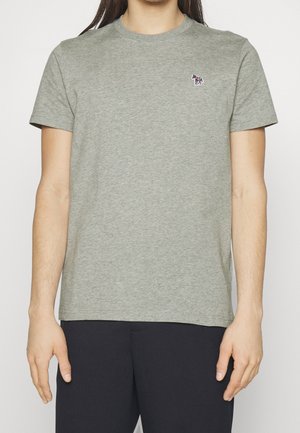 Osoba nosząca prosty szary t-shirt z krótkim rękawem, z małym kolorowym logo zebry na górnej lewej stronie klatki piersiowej, w połączeniu z czarnymi spodniami.