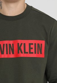 Felpa verde scuro con un logo rettangolare rosso che presenta la scritta "CALVIN KLEIN" in grassetto nero. Design a girocollo.