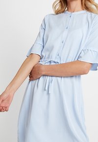 Jasnoniebieska, koszulowa sukienka z krótkimi, falbaniastymi rękawami i talią regulowaną sznurkiem. Gładki materiał o luźnym kroju, sięgający do połowy łydki.