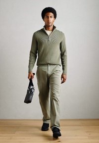 Grøn stribet mønster zippered sweater, lysebeige bukser, sorte sko, holder en lille sort taske med rem. Trægulv som baggrund.