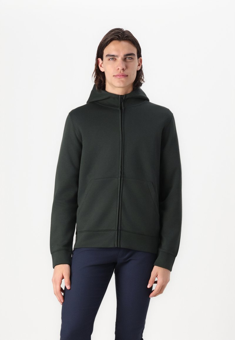 Ciemnozielona bluza zip bezrękawnik wykonana z miękkiego materiału, z kapturem, przednimi kieszeniami i dopasowanymi mankietami. Model ma na sobie granatowe spodnie.