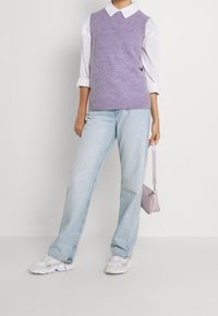 Fioletowy sweter bezrękawnik na białej koszuli z kołnierzykiem, w zestawie z jasnoniebieskimi dżinsami i białymi sneakersami. Trzyma małą lawendową torebkę.