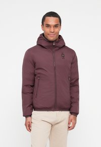 Chaqueta con capucha burdeos con cierre de cremallera, dos bolsillos laterales y una superficie texturizada. Presenta un pequeño parche de logo en el pecho.