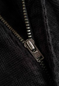 Schwarzer Reißverschluss mit metallischen Zähnen und Ziehlasche auf strukturiertem Stoff. Nahaufnahme, die das Design und die Verarbeitungsqualität des Reißverschlusses hervorhebt.