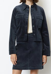 Tmavě modrá bunda ze směsi bavlny s límcem, dvěma kapsami na prsou a zapínáním na knoflíky, doplněná o odpovídající úzkou sukni. Hladká textura.