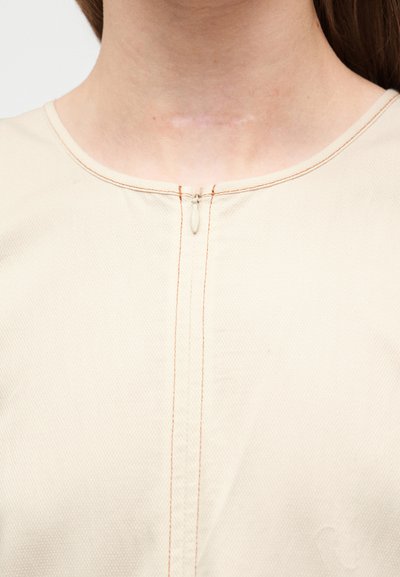 Μπεζ υφασμάτινο ένδυμα με στρογγυλή λαιμόκοψη, κεντρικό φερμουάρ και πορτοκαλί ραφές σε αντίθεση. Λείο ύφασμα.