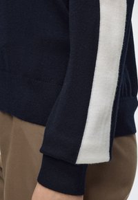 Manche de pull bleu marine avec une bande verticale blanche portée sur un pantalon marron, montrant un poignet côtelé et un tissu tricoté texturé.