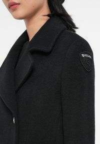 Manteau en laine noir avec un large col et une finition texturée. Une patch en forme de bouclier noir avec le nom de la marque "Blauer" y est attaché.