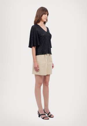 Black, sparkly V-neck top with flared sleeves, paired with beige shorts. Model stands sideways, wearing black strappy sandals.