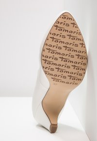 Chaussure à talon haut blanche avec un bout pointu et un matériau lisse. La semelle extérieure présente le nom de la marque "Tamaris" dans un motif répétitif. Taille 37.