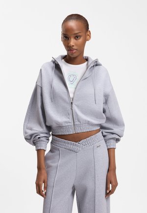 Woman wearing light gray cropped zip-up hoodie with puffed sleeves and matching high-waisted pants with a v-shaped waistband.