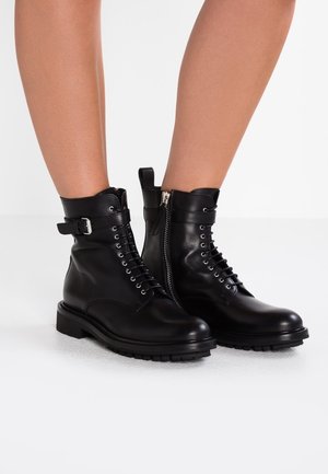 Czarne skórzane botki za kostkę z wiązaniem z przodu, zamkiem błyskawicznym z boku i paskiem z klamrą. Charakteryzują się masywną, bieżnikową podeszwą i gładką teksturą.