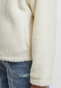 Cremefarvet fleecejakke med en tekstureret overflade, der har en brystlomme og en blød, afrundet kanten, båret over slidte denim. 