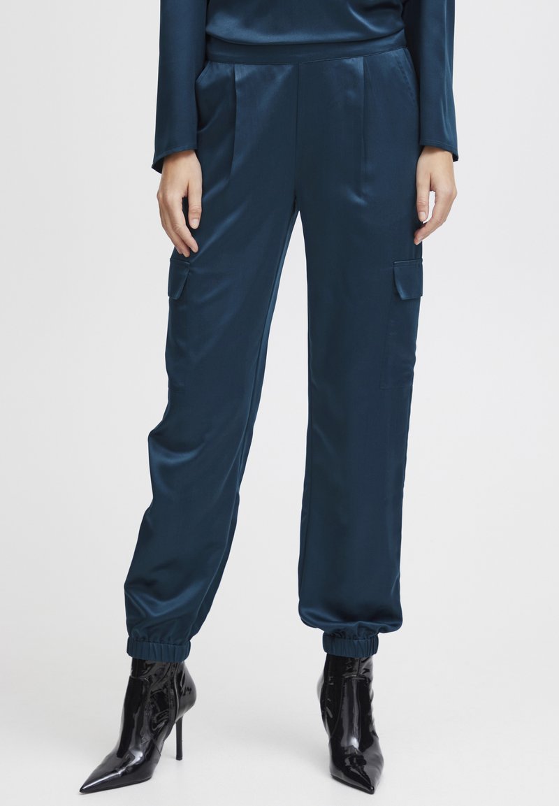 Pantaloni cargo din satin într-o nuanță închisă de teal, cu manșete elastice și buzunare laterale, asortați cu cizme negre cu vârful ascuțit.