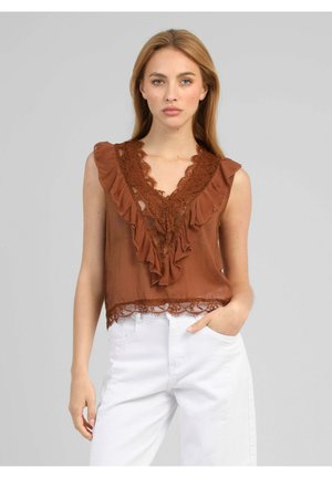 Femme portant un chemisier marron sans manches en dentelle avec volants au col en V et un pantalon blanc, debout devant un fond gris uni.