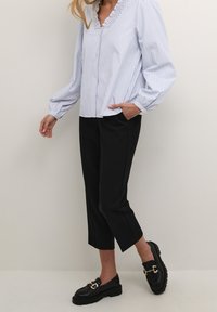 Blouse rayée bleue et blanche avec un col volant, manches longues, pantalons noirs cropped et mocassins noirs épais avec des accents dorés.