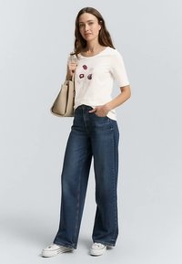 Weißes Kurzarmhemd mit Blumenstickerei, kombiniert mit hoch taillierter, weiter blauer Jeans und weißen Sneakern. Beigefarbene Handtasche inklusive.