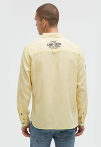 Gelbes Langarmhemd mit einem Logo auf der Rückseite, geknöpften Manschetten und einem abgerundeten Saum. Der Stoff scheint strukturiert; die Farbe ist weich und gedämpft.