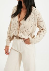 Knit beige cardigan with diamond pattern and small bobble accents, featuring a deep V-neck and button closure, paired with cream high-waisted pants.