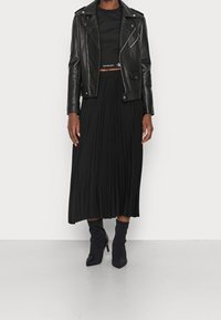 Chaqueta de motorista de cuero negro sobre un top negro corto, combinada con una falda midi negra con pliegues y botas negras hasta el tobillo, con puntas puntiagudas.
