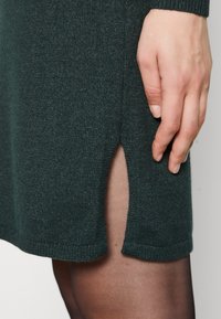 Abito lavorato a maglia verde scuro con spacco laterale e orlo a coste, abbinato a collant neri trasparenti. Mano posizionata vicino allo spacco per una migliore visibilità dei dettagli.