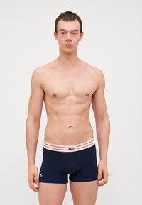 Marineblauwe boxershorts met een aansluitend ontwerp, voorzien van een witte waistband met rode strepen en een klein groen logo aan de zijkant.