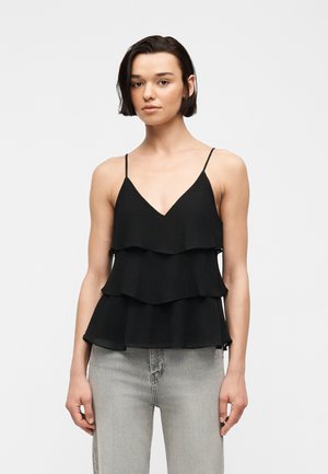 Ung kvinde med kort mørkt hår iført en sort lagdelt camisole-top og lysegrå jeans, stående mod en ensfarvet baggrund.
