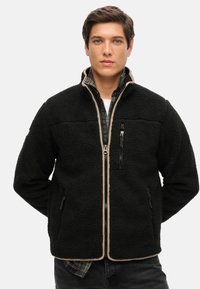 Giacca in pile nera con zip davanti, dettagli beige e tasche laterali. Materiale testurizzato e colletto alto. Indossata sopra una camicia a scacchi.