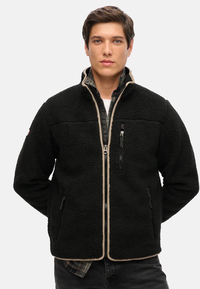 Giacca in pile nera con zip davanti, dettagli beige e tasche laterali. Materiale testurizzato e colletto alto. Indossata sopra una camicia a scacchi.