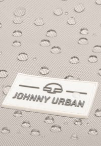 Gouttes d'eau sur un tissu texturé gris clair avec une étiquette rectangulaire blanche affichant le logo en relief noir "Johnny Urban".