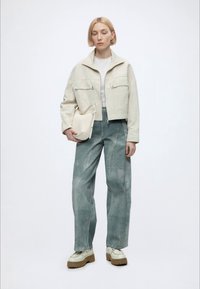 Femme blonde portant une veste beige courte, une chemise blanche, un jean large bleu clair, des chaussures beige épaisses, tenant un sac à main beige.
