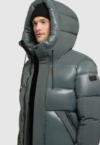 Grå pufferjakke med stor hætte, glat overflade, quiltet design, frontlynlås og sidelommer med sorte detaljer og snore.