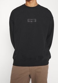 Felpa nera realizzata in tessuto morbido, con scollatura a girocollo e spalle scese. Presenta un logo grigio Wrangler sul petto.