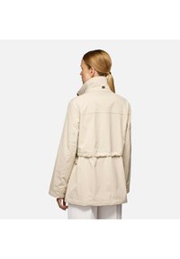 Giacca beige con colletto alto e vita regolabile. Presenta una texture liscia e un logo discreto sulla schiena, pensata per una vestibilità comoda.