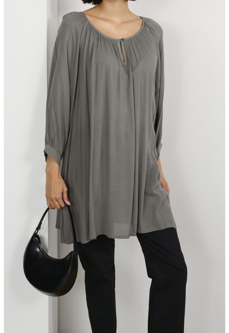 Femme portant une tunique grise ample à manches longues avec détail de boutons, pantalon noir, tenant un petit sac à main noir sur fond blanc.