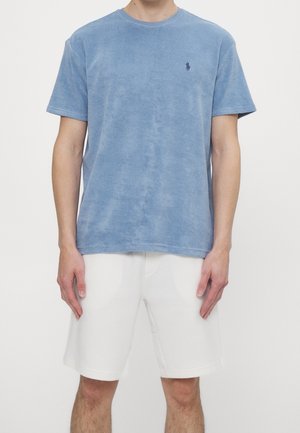 Man draagt een lichtblauw shirt met korte mouwen en een klein logo op de borst, en witte shorts tot aan de knieën, staande tegen een effen witte achtergrond.