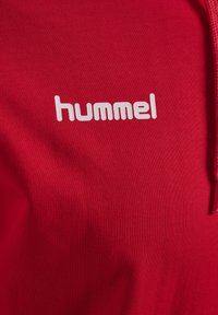 Roter Stoff mit weiß gedrucktem "hummel"-Logo, der einen Teil eines Kordelzugs auf der rechten Seite zeigt.