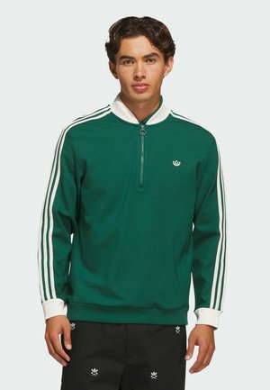 Joven con camisa deportiva verde de manga larga con cremallera, rayas blancas en las mangas y cuello blanco, de pie sobre un fondo liso.