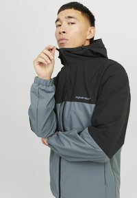 Herrejakke med et to-tonet design, der har en sort overdel og en grå underdel. Materialet er glat med en justerbar hætte og lynlås lukning.